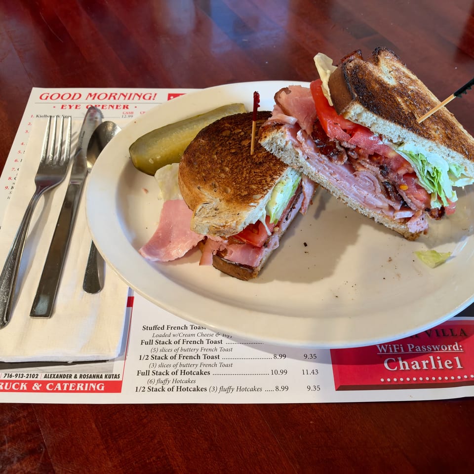 Toasted bacon, lettuce, tomato, and ham sandwich with pickle spear on a plate at a casual diner.