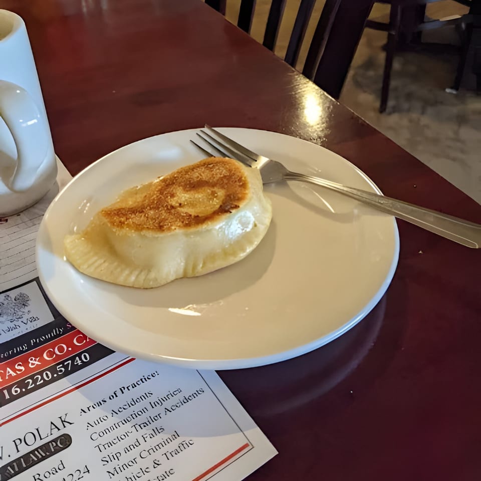 A golden-brown pierogi on a white plate with a fork, next to a white mug and a paper.