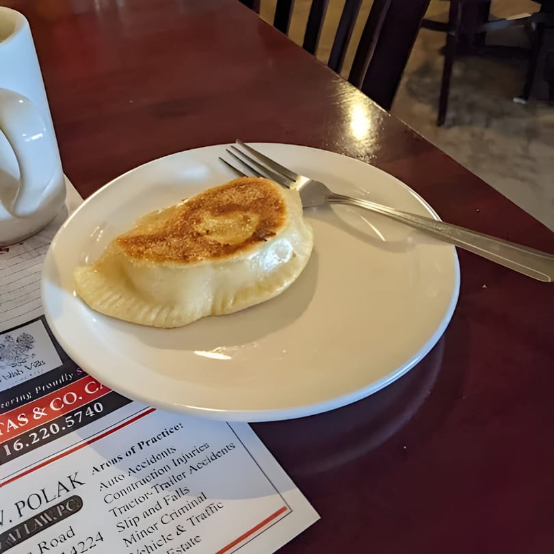 A golden-brown pierogi rests on a white plate with a fork, on a dark red table.